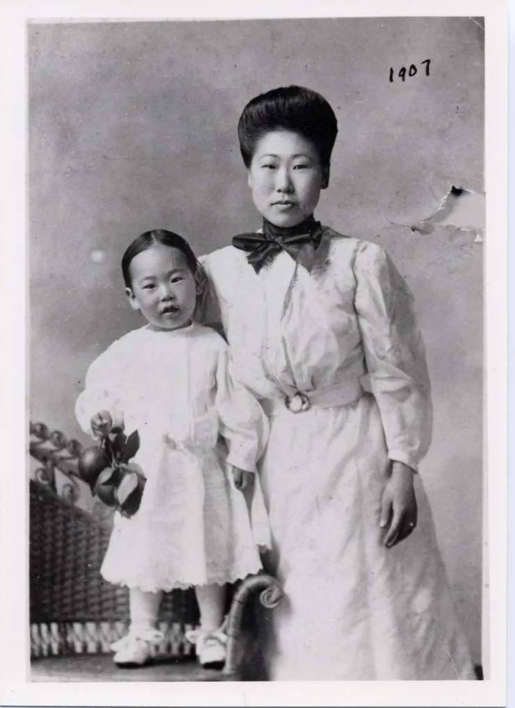 A studio photograph of Yi Hye Ryon (Helen Ahn) standing in a long white dress holding a small child, 1907
