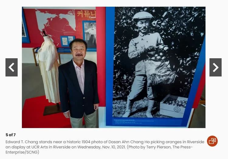 A man in a dark jacket standing in a gallery beside a wall-height reproduction of the orange-picking photograph