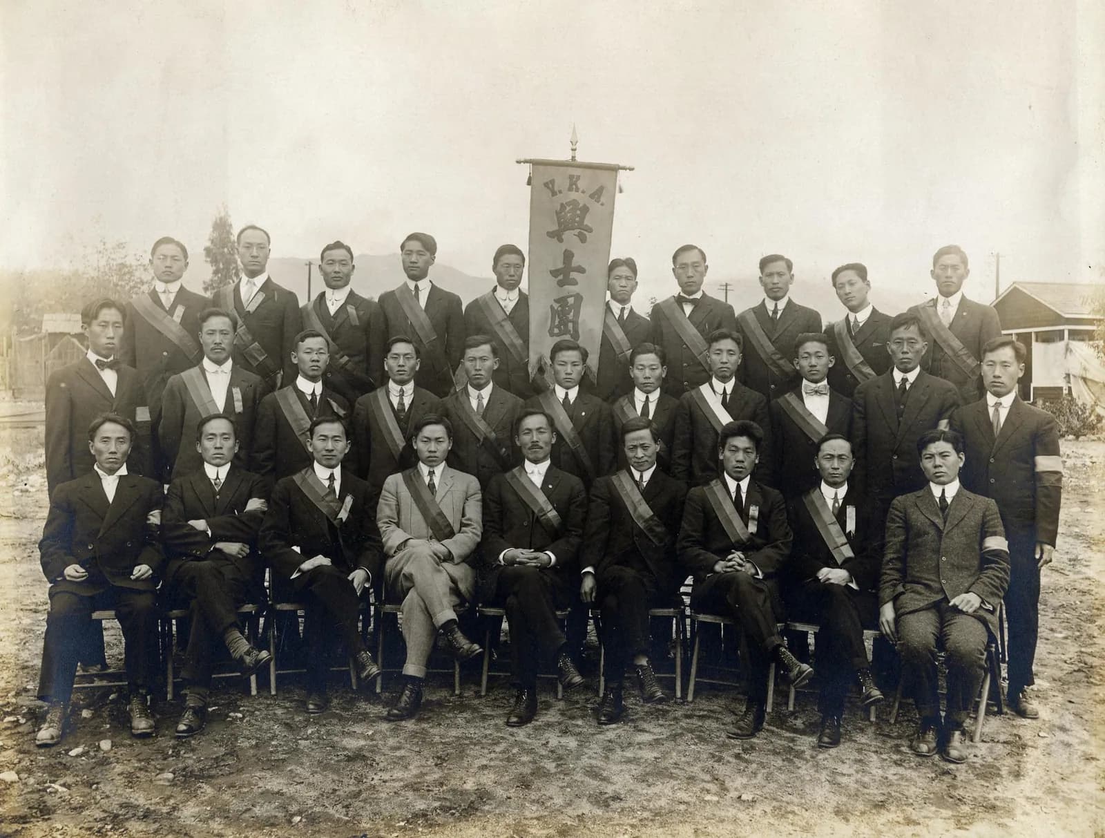 About thirty men in suits, most wearing sashes, in three rows around a standing banner lettered Y.K.A. and 興士團