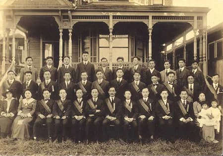 Rows of men in dark suits and women in white hanbok posed in front of a wooden porch