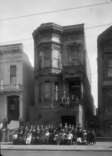 A tall Victorian building with bay windows, a group of people gathered on its front steps and the pavement below
