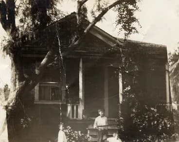 A wooden house half hidden by a tree, small figures on its porch